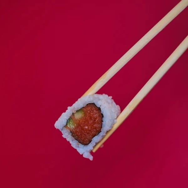 Chopsticks holding a single sushi roll with red filling and white rice against a red background.
