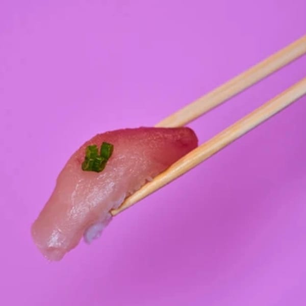 Chopsticks holding a piece of Yanagi Kitchen sushi with red tuna and green garnish on a purple background.
