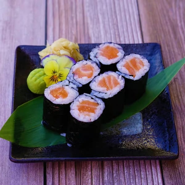 Six salmon sushi rolls with ginger, wasabi, and a pansy on a dark plate.