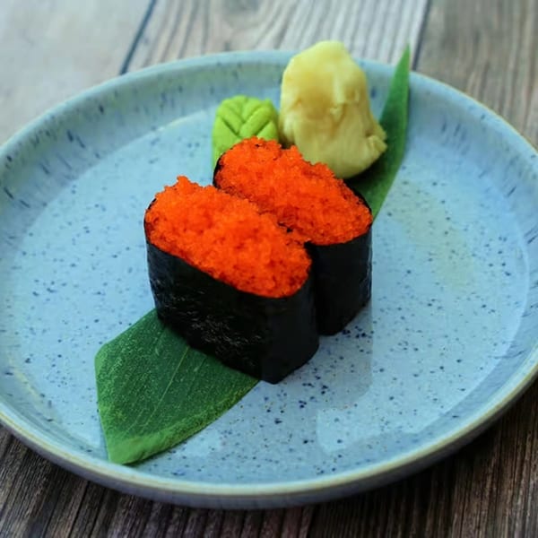 Two gunkan sushi pieces with bright orange tobiko, wasabi, and ginger on a blue plate.