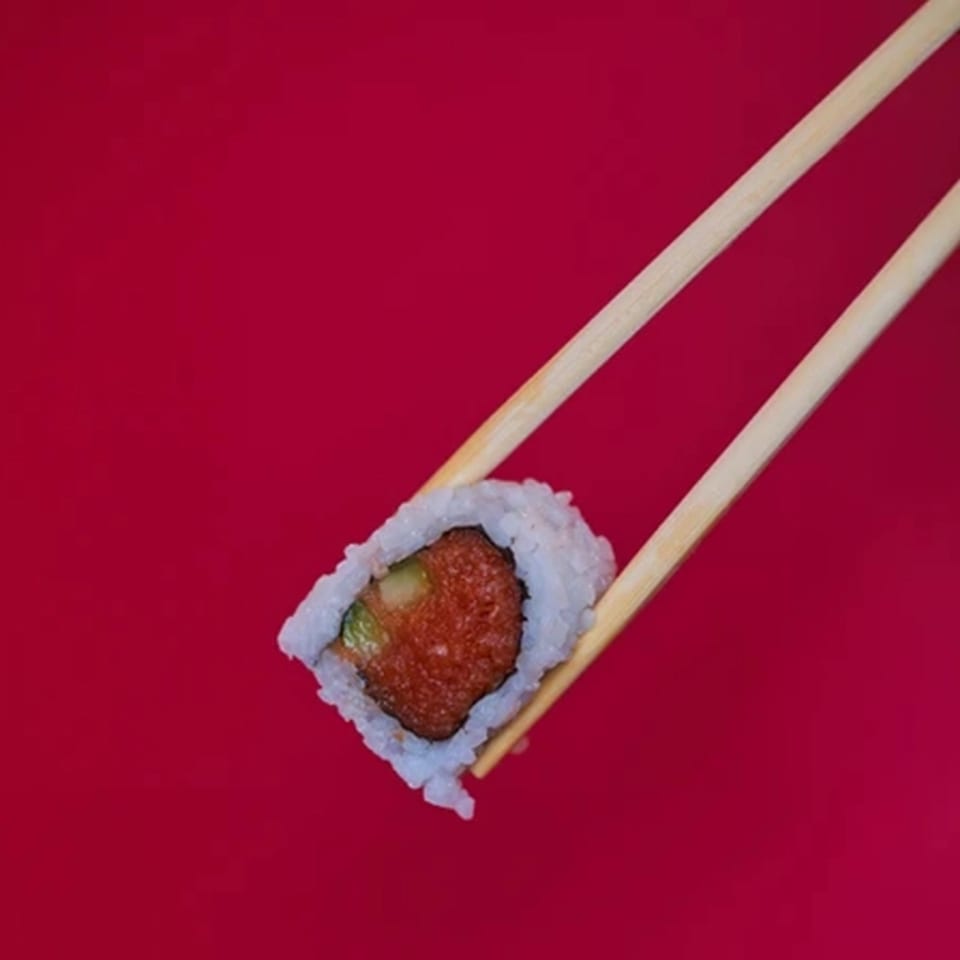 Chopsticks holding a single sushi roll with red filling and white rice against a red background.