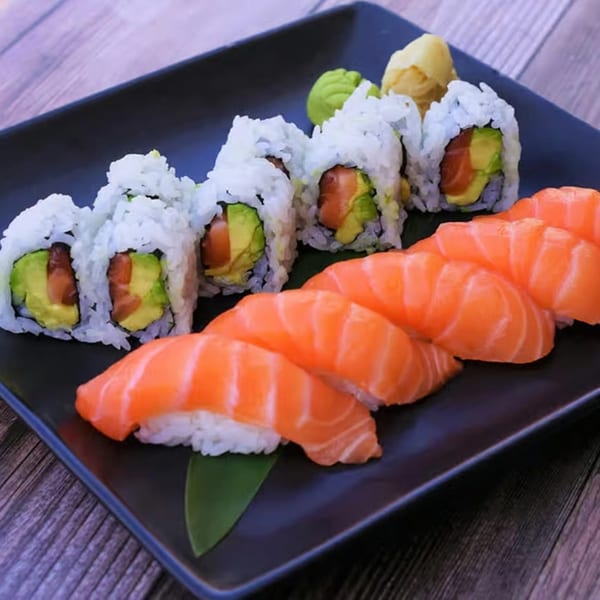 A black rectangular plate holds salmon nigiri, California rolls, wasabi, and pickled ginger on a wooden table.